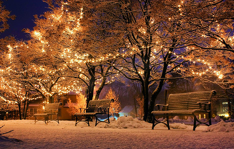 雪下灯笼摄影照片_圣诞树下的长凳和雪中闪亮的灯笼的景色。