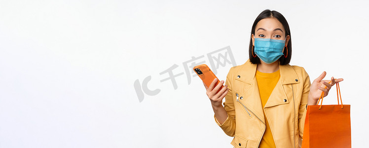时尚亚洲女购物者的形象，手持智能手机和没有商店标志的购物袋，戴着 covid-19 的医用面罩，白色背景
