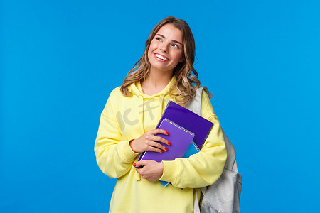 梦幻快乐、漂亮的金发女孩在大学学习，拿着背包和笔记本，家庭作业人员，看起来左上角深思熟虑，站在蓝色背景
