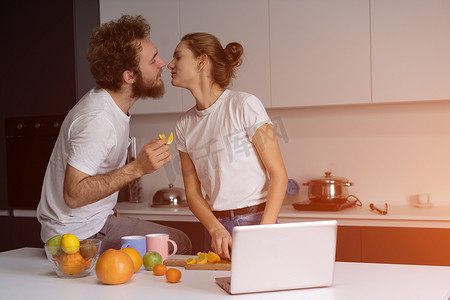 美丽的年轻夫妇亲吻互相喂食，在现代厨房里取笑，在家里做饭时微笑。