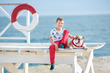 英俊的男孩青少年快乐地与他的朋友斗牛犬在海边度过时光孩子狗抱着玩两个海星靠近救生圈漂浮穿着红色裤子裤子拖鞋和 T 恤