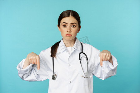 悲伤而阴郁的医生，女医务工作者手指向下，看起来很沮丧，显示坏消息，站在蓝色背景的白大衣上
