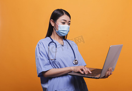 年轻的成年护士在使用笔记本电脑时戴着病毒防护面罩和听诊器。