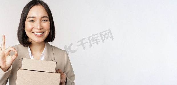 微笑的亚洲女商人，展示好的标志和装有送货的盒子，为客户准备订单，站在白色背景上