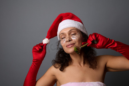 穿着圣诞老人服装的漂亮女人使用玉石滚轮按摩器进行整容和淋巴引流面部按摩。
