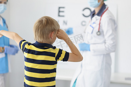 眼科诊断摄影照片_男孩在诊所、眼科柜、视力诊断闭眼并在船上大声说出字母