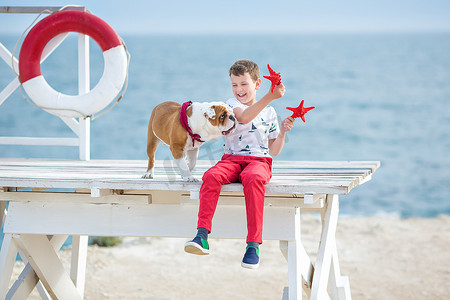 英俊的男孩青少年快乐地与他的朋友斗牛犬在海边度过时光孩子狗抱着玩两个海星靠近救生圈漂浮穿着红色裤子裤子拖鞋和 T 恤