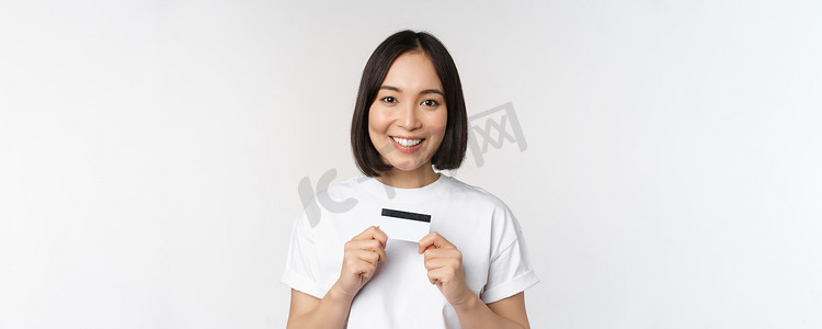 微笑的韩国女性展示信用卡，脸上挂着幸福的表情，站在白色背景的 T 恤上