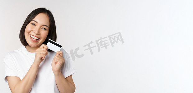 银行产品摄影照片_微笑的亚洲女性拥抱信用卡、购买非接触式产品、站在白色背景上的白色 T 恤的形象