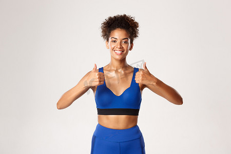 美丽的非洲裔美国健身女性腰部以上，穿着蓝色运动服，竖起大拇指表示赞同，推荐锻炼装备，白色背景