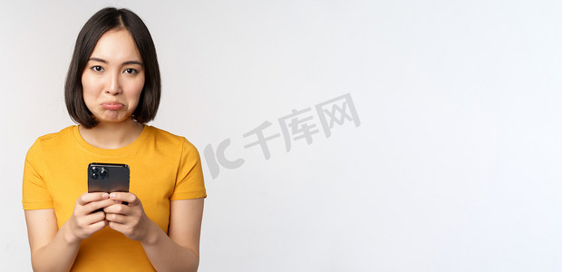 悲伤的亚洲女性拿着智能手机，看上去懊恼不已，站在白色背景下的黄色 T 恤上