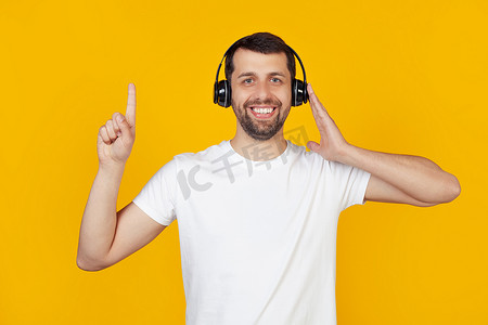 穿着白色 T 恤、留着胡子的年轻人在孤立的黄色背景下戴着耳机听音乐，对一个想法或问题感到惊讶，用手指指着一张快乐的脸，第一