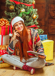穿着冬季针织毛衣、帽子和条纹袜子的美丽女人坐在地板上，双腿交叉，靠近一堆礼物和一棵圣诞树。
