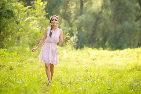 浅色花环摄影照片_年轻漂亮的女人穿着浅色夏装，在绿色的草地上行走，手里拿着一束野花