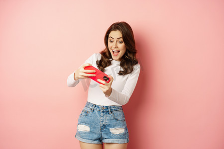 玩手机视频游戏的快乐美女，水平拿着智能手机，用兴奋的脸，粉红色背景在手机上观看