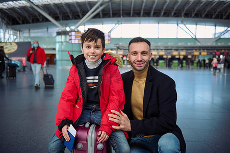 红色羽绒服摄影照片_白人男子 — 快乐的父亲和可爱的十几岁儿子穿着红色羽绒服坐在手提箱上拿着护照，登机牌在国际机场出发区微笑着看着相机