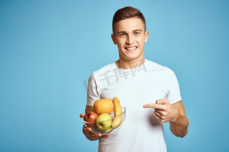 香蕉橙子摄影照片_带新鲜水果的快乐男人用手示意蓝色背景白色 T 恤维生素香蕉橙子苹果