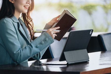 一位女企业家或女商人在阅读一本制定金融和投资策略的书并操作在木桌上工作的平板电脑时面带微笑。