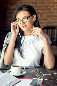 手机听音乐摄影照片_戴眼镜的美丽微笑女人正在咖啡馆用智能手机听音乐。
