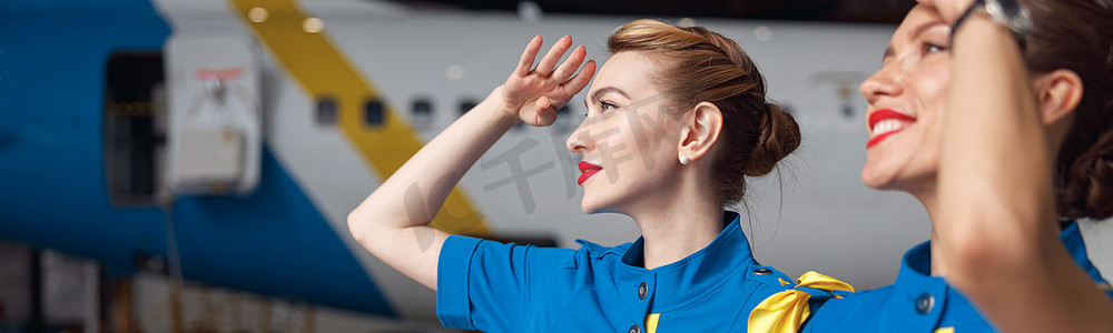 漂亮空姐摄影照片_两位身穿时尚蓝色制服的美丽空姐的肖像，微笑着仰望天空，一起站在机场机库的客机前