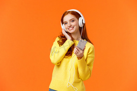 冬天的音乐背景摄影照片_腰上画像开朗可爱的年轻女性，红头发，戴着耳机听音乐，倾斜头，满意地微笑，拿着智能手机，为温柔平静的夜晚挑选歌曲，橙色背景