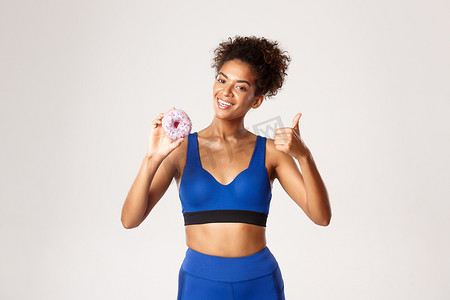 身着蓝色运动服、面带微笑的非洲裔美国女运动员的腰部向上拍摄，竖起大拇指和甜甜圈，微笑着高兴，白色背景