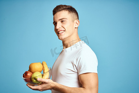 带新鲜水果的快乐男人用手示意蓝色背景白色 T 恤维生素香蕉橙子苹果
