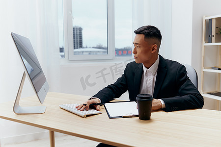 身穿西装、咖啡杯的亚洲男子坐在电脑前的桌子旁