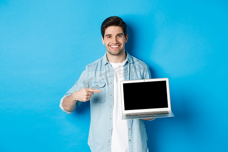 英俊的年轻人指着电脑屏幕，微笑着高兴，在互联网或网站上展示促销，站在蓝色背景上