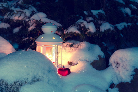 雪下灯笼摄影照片_在雪堆的房子庭院里，一棵被雪覆盖的圣诞树下，有燃烧的蜡烛的灯笼