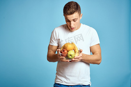 带新鲜水果的快乐男人用手示意蓝色背景白色 T 恤维生素香蕉橙子苹果