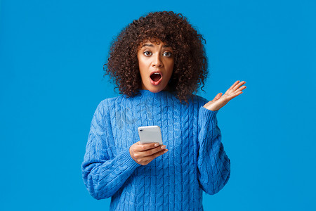 忧心忡忡、震惊的年轻非洲裔美国女性通过智能手机收到不愉快的消息，用优柔寡断的表情告诉它，耸耸肩，举起手，不知道该做什么，蓝色背景