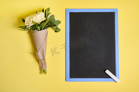 一束精致的秋季紫苑花的平铺组合物，用绳子绑在工艺包装纸上，空白的空黑板在黄色背景上有文字空间