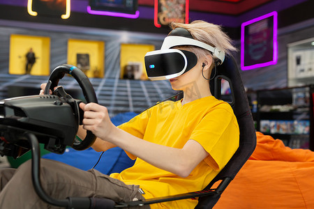 戴着虚拟现实眼镜的十几岁男孩握着方向盘，在控制台上玩电脑游戏。