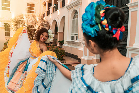 两名年轻的拉丁裔女性，一名混血儿和一名西班牙裔，在殖民时期的房屋外穿着尼加拉瓜的传统服装跳舞