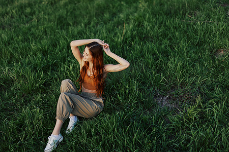 红色长围裙摄影照片_夏天，一位长着红色卷发的漂亮女人坐在公园里，夕阳下的绿草上