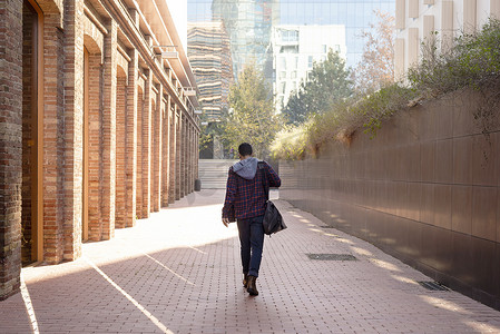 一个迷人的年轻人拿着肩包穿过城市的背面图
