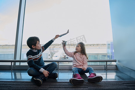 坐飞机的儿童摄影照片_小男孩和小女孩，弟弟妹妹坐在机场出发航站楼全景窗户旁的地板上玩玩具飞机，可以俯瞰跑道。