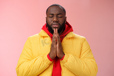 穿着黄色外套的严肃忠实的年轻坚定的非洲裔美国人祈祷者双手合十祈祷闭上眼睛耳语恳求上帝帮助许愿，站立粉红色背景