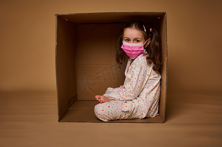 穿着睡衣、戴着粉色医用防护面具、扎着两条马尾辫的可爱欧洲小女孩坐在纸箱里，看着相机，在米色背景中被隔离，上面有复制广告空间
