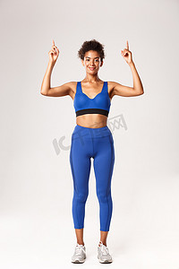 穿着蓝色运动服、面带微笑、迷人的黑人女运动员，看着镜头很高兴，手指向上，展示关于锻炼的标志或广告，白色背景