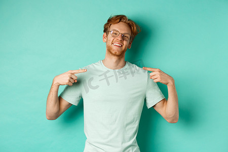 自信的红头发男人戴着眼镜和 T 恤，面带自鸣得意的微笑，指着自己，站在绿松石背景上吹牛