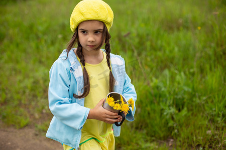 春天黄色蒲公英摄影照片_一个小女孩走在绿色草坪附近的一条小路上，手里拿着一束黄色蒲公英