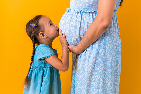 母亲、爱情、童年、怀孕、炎热的夏天 — 裁剪肖像怀孕的无法辨认的母亲蓝色连衣裙小学龄前女儿女孩兄弟姐妹泰迪熊拥抱黄色背景的妈妈肚子