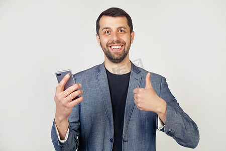 手势很棒的标志摄影照片_微笑的年轻商人，一个穿着夹克、留着胡子的男人，手里拿着智能手机，开心地笑着，做出一个好的手势，用手指竖起大拇指，这是一个很好的标志。