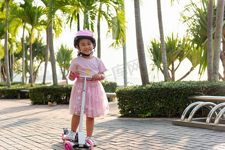 夏日，快乐的亚洲小女孩戴着安全头盔，在户外公园的路上玩粉色踢板
