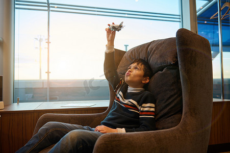 坐飞机的儿童摄影照片_帅气的白人青春期男孩坐在椅子上玩玩具飞机，全景窗户旁俯瞰日落时分的跑道，同时在机场出发航站楼等待登机