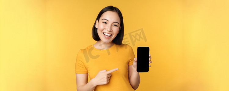 美丽快乐的亚洲女孩展示手机屏幕，在智能手机小工具上的应用，站在黄色背景上