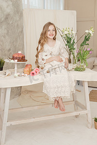 女孩坐在复活节餐桌旁，手里拿着蛋糕、春花和鹌鹑蛋，怀里抱着一个白人山羊。