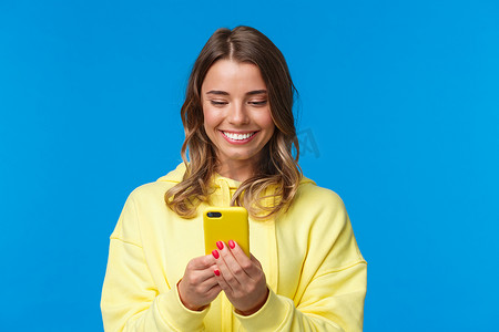 特写肖像愚蠢美丽的金发欧洲女人，穿着黄色连帽衫，在手机显示屏上微笑，看着可爱的视频，与男友发短信，站在蓝色背景